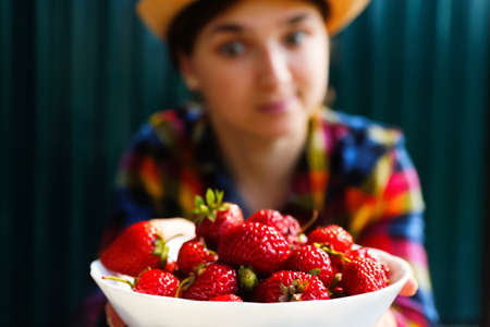 Defocus young woman in a hat and shirt smiles and shows a white bowl of strawberries. Dark green steel background. Summer food, fruits. Summertime. Healthy lifestyle. Diet concept. Out of focusの写真素材