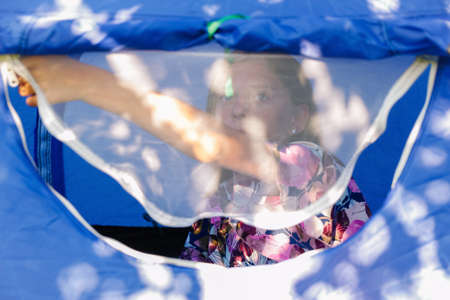 The girl sitting comfortably inside a camping blue tent behind a mesh anti-mosquito window, she looking outside. Portrait teen girl. Out of focus.の写真素材