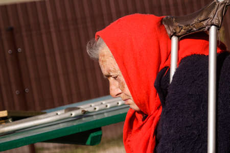 Defocus ukrainian elderly woman in red shawl with crutches sitting outside. Woman 85 years old. Alone person. Closeup face. Sick and homecare. Sad people. Dementia. Copy space. Out of focus.の写真素材
