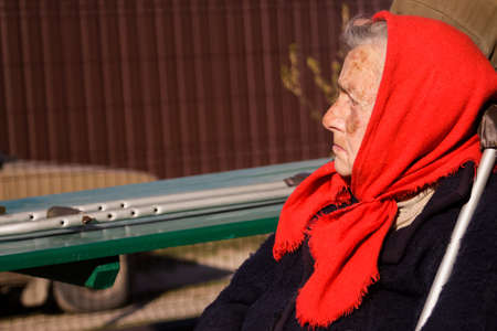 Defocus ukrainian elderly woman in red shawl with crutches sitting outside. Woman 90 years old. Alone person. Closeup portrait. Sick and homecare. Out of focusの写真素材
