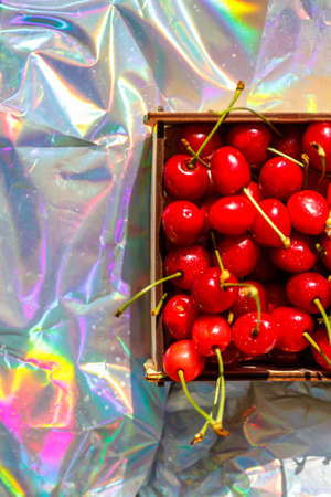 Defocus close-up cropped box, crape of dark red wet sweet cherries with tail and leaves on modern hologram background. Fruits and berries. Harvest and crop concept. Organic, eco. Out of focusの写真素材