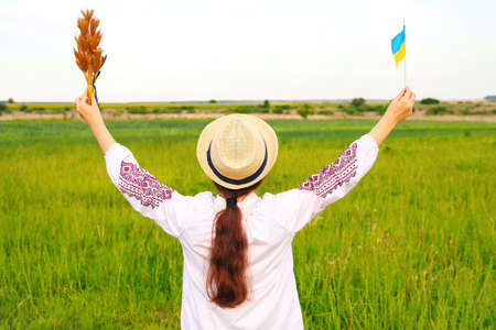 Defocus young woman in vyshyvanka and hat holding bouquet of ripe golden spikelets of wheat tied and...