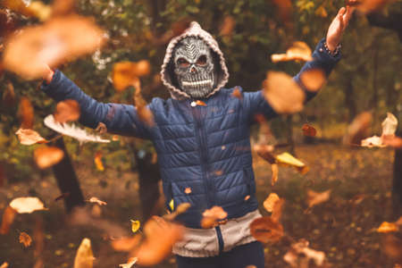 Defocus Halloween people. Person in grim reaper mask raising hand and throwing leaves. Many flying orange, yellow, green dry leaves. Skull ghost. Fall park. Out of focus.の写真素材