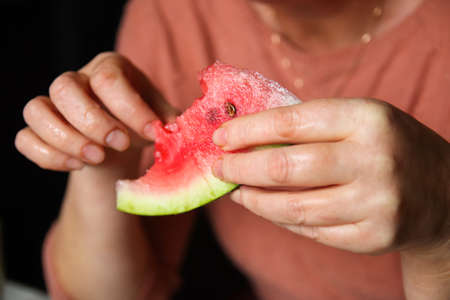 Defocus woman eating watermelon. Woman's hand holding slice of watermelon in home. Closeup female hands holding red fruit. Out of focus.の写真素材
