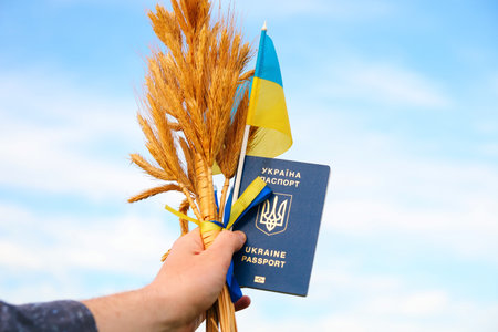 Defocus young man holding ukrainian passport, flag and wheat spikes of wheat tied and flag on the blue sky background. Flag Ukraine. Freedom. Ukrainian men. Closeup. Freedom. Out of focus.の写真素材