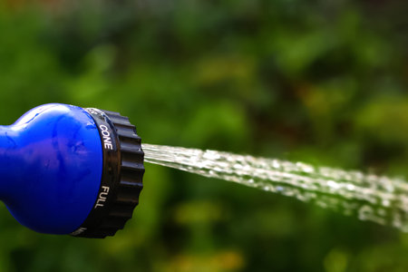 Defocus closeup water garden. Female hand holding blue automatic sprinkler sprays water over brown blurred background. Gardener watering the lawn in the garden. Out of focus.の写真素材