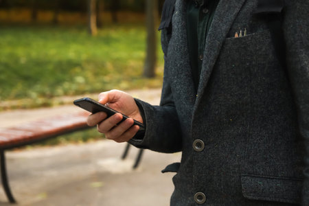 Man phone. Defocus male hand holding phone. Using smart phone in autumn park. Typing text message or reading social media at mobile phone. Fall background. Cell phone. Communication. Out of focus.の写真素材
