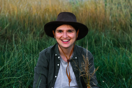Smiling relaxed woman breathing fresh air in a green meadow. Girl in a cowboy hat in a field. Sunset. Nature background. Closeup portrait caucasian girl. Summertime. Wellness. Out of focus.の写真素材