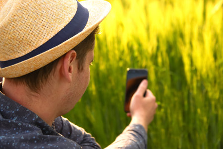 Man with phone on sunset. Young farmer working in a wheat field, inspecting and tuning irrigation center pivot sprinkler system on smartphone. Out of focus.の写真素材