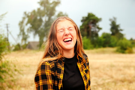Smiling happy girl laughing emotions. Portrait preteen girl on the nature background. Girl 14 years old. Blonde long hair. Attractive teenager. Smiling teen girl. Closed eyes. Out of focus.の写真素材