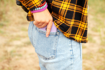 Woman puts mobile phone in jeans pocket on yellow nature background. Blue denim. Smart phone in the purple case. Mobile call. Out of focus.の写真素材