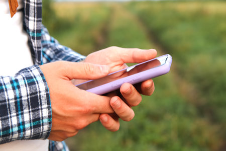 A close-up shot captures a male Caucasian farmer's hands firmly grasping a smartphone in an outdoor setting.の写真素材