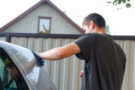A man washing a gray car with a sponge at a car wash. He holds a microfiber cloth and is polishing...