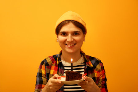 Young Generation Z woman celebrating her birthday with a cake topped with a lit candle. She wears...