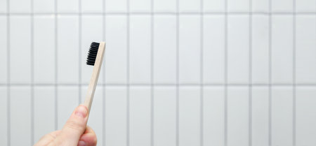 A wooden eco-friendly toothbrush held in a hand against a clean white tile background, emphasizing...