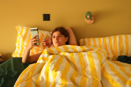 A candid image of a woman in bed in the morning, mindlessly scrolling through social media on her phone. social media addiction woman bed. digital detox phone mental health. device dependency.の写真素材