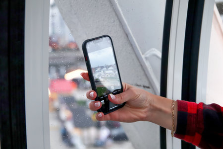 a female hand holding a phone, takeing a shot of a city landscape in the rain. The photo, a powerful...