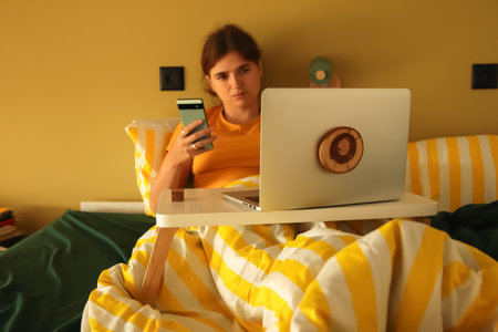 digital distraction smartphone bed yellow background. remote work distraction woman laptop phone. A woman is lying on a bed surrounded by her phone smartphone and laptop, avoiding work.の写真素材