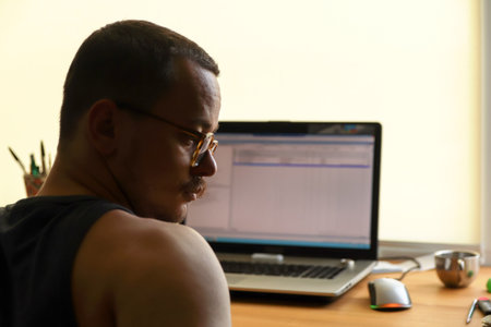 A portrait of a man in profile, seated with his laptop. His thoughtful expression, highlighted by his eyewear and moustache, hints at procrastination or deep, focused thought while working.の写真素材