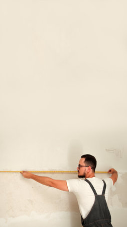A focused, vertical image of a construction worker in an overall uniform, precisely taking measures of a clean, white wall at a construction site. worker taking measures white wall construction.の写真素材