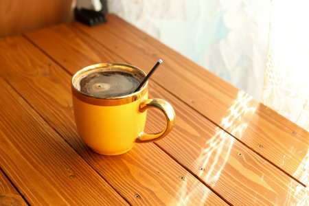 A yellow ceramic cup with a gold rim filled with black coffee and a spoon, sitting on a wooden table in bright morning sunlight.の写真素材