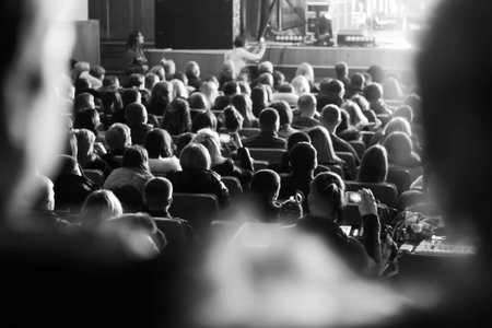 Black and white shot of an audience sitting in a theater or concert hall, looking at a brightly lit stage. A person in the foreground is filming the performance on a smartphone.の写真素材