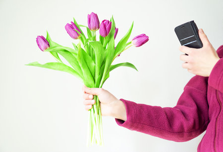 Beautiful girl in cozy purple jacket with magenta tulips is taking a picture on her phone.の写真素材