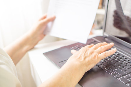 Close up of middle aged female hands with laptop and paper document, working and tipping on keyboardの写真素材