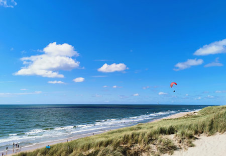Paraglider flying above a sandy beach and coastal dunes along the sea under a bright blue sky with...