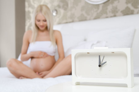young pregnant woman sit in light  home room, focus on clockの写真素材