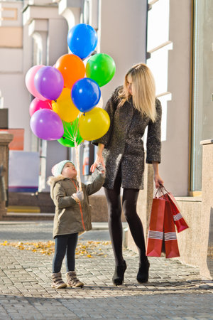 happy woman and little child with red shopping bag and air-balloons, walking on street