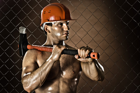 the muscular  tired worker chopper man, in  safety helmet  with big  heavy ax  in hands, on netting fence backgroundの写真素材