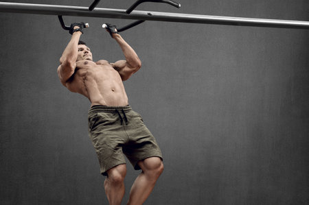 Guy bodybuilder perform exercise do chin-ups, horizontal bar on gray wall background with empty...