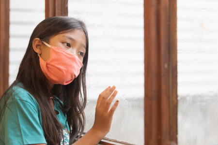 An Asian elementary school girl wearing a mask is in a sad mood at the window.. The picture shows...