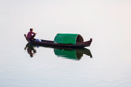 Mandalay/Myanmar-October 6th 2019: A Burmese fisherman sits on a wooden fishing boat and uses a net to trap fish in the lake.のeditorial素材