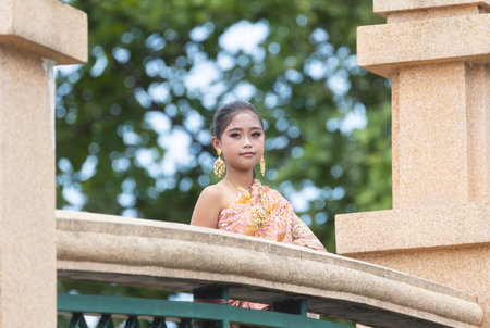 An Asian schoolgirl dressed in traditional Thai clothes stands at the railing of a bridge in a park.の写真素材