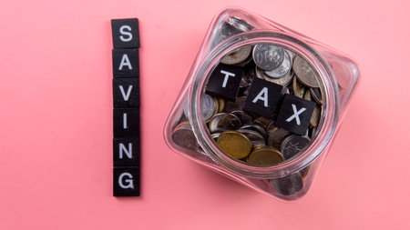 Saving tax payment concept. Selective focus of a word tile, fake money and a jar of coins on isolated on pink backgroundの写真素材