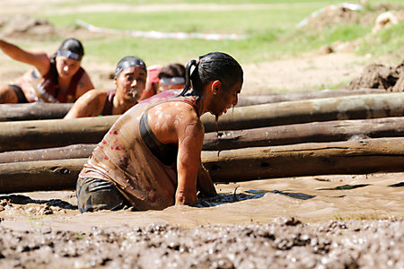 Female athlete crawling through muddy water obstacle during obstacle course race, with other...