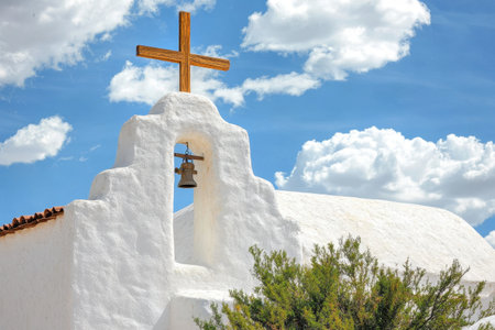 A historic church features a wooden cross atop its whitewashed structure, complemented by a bell. Fluffy clouds drift in a vivid blue sky, enhancing the peaceful atmosphere.の素材