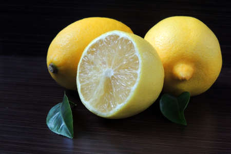 Macro view of vibrant, yellow lemon slice and two whole lemons on dark brown, rustic, wooden table,...