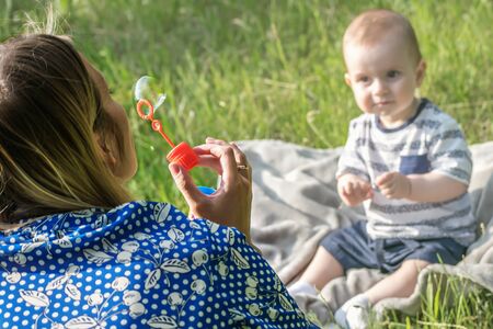 Young mather inflating soap bubbles for Surprised baby boy sitting on grassの写真素材