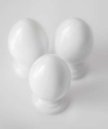 Top view of three white decorative ceramic eggs on stands or in egg-cups on white blurred background. Easter religious Christian symbol. Monochrome imageの写真素材