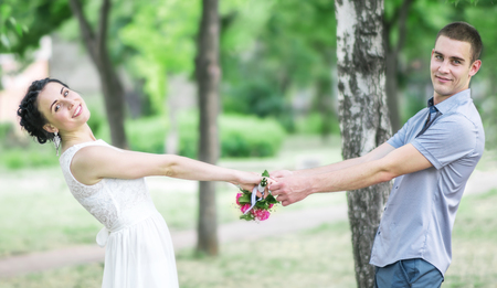 Portrait of happy beautiful young couple female bride with small wedding flowers roses bouquet and male bridegroom holding hands, smiling and spinning in summer parkの写真素材