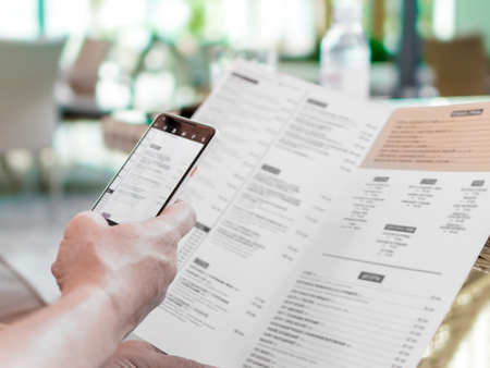 Person's hands holding menu and mobile phone in restaurant. A foreign man in cafe ordering food using word translation program while sitting at table. Mockup for smartphone screen.の写真素材