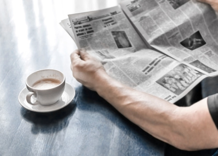 A man reading newspaper and drinking coffee sitting in cafe or at home in kitchen at morning time. A coffee cup on dark blue table. A person hands holding newspaper.の写真素材
