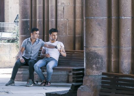 CYPRUS, NICOSIA - JUNE 10, 2019: Two male sitting and talking together on a bench near the ancient columns of the historic building Selimiye Mosque. One young man holding a smartphoneのeditorial素材