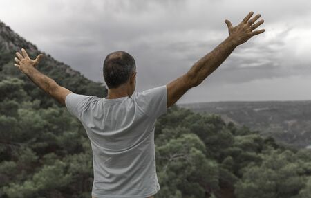 An elderly man standing on top of a mountain with his arms outstretched. Beautiful view of the mountains and sky from the back of a man.の写真素材