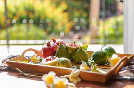 Fresh whole and cut fruits figs, pomegranate and exotic plumeria flowers on wooden tray on table next to open window background. Still life.の写真素材