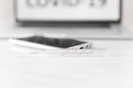 Laptop with the inscription Covid-19, medical thermometer and smartphone lying on white table. World coronavirus Pandemic 2020. Background with copyspace.の写真素材