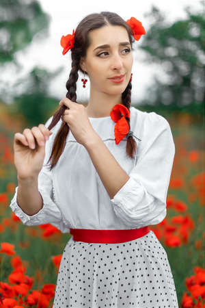 Young pretty girl Slavic or Ukrainian posing in folk dress on a flowering poppy field. Female weaving braid.の写真素材